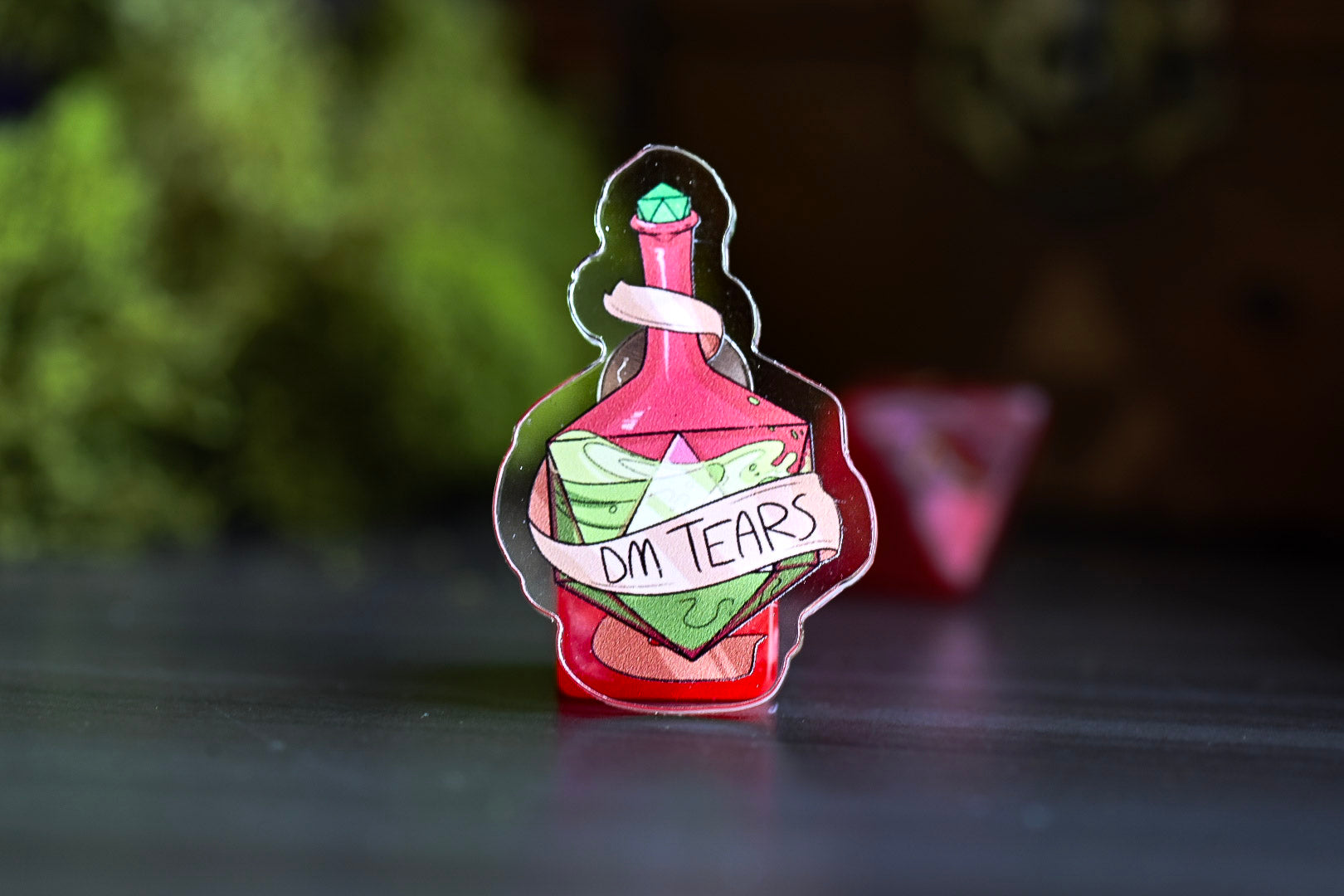 Enamel pin of a potion bottle labeled 'DM Tears' on a dark surface with a blurred green and brown background.