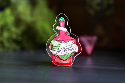 Enamel pin of a potion bottle labeled 'DM Tears' on a dark surface with a blurred green and brown background.